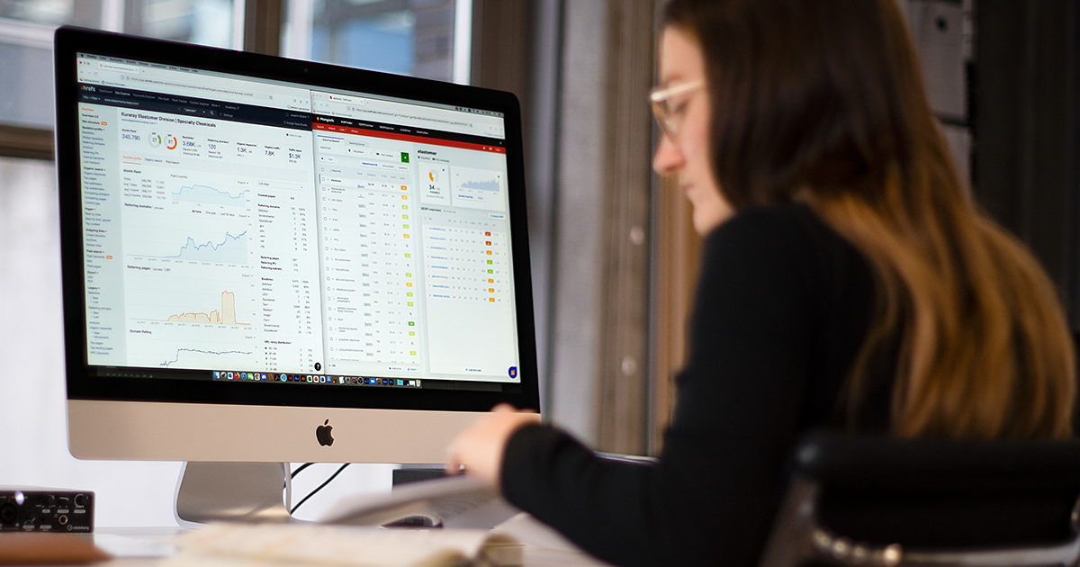 Man sieht eine junge Frau vor einem Computer sitzen. Auf dem Bildschirm sieht man zwei Programme zur Keyword Recherche.